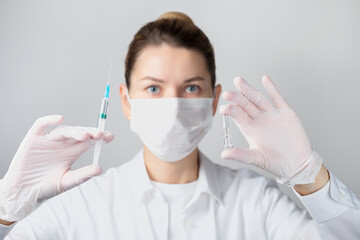 Close-up of a woman in a protective face mask with a syringe and an ampoule in her hand on a gray...