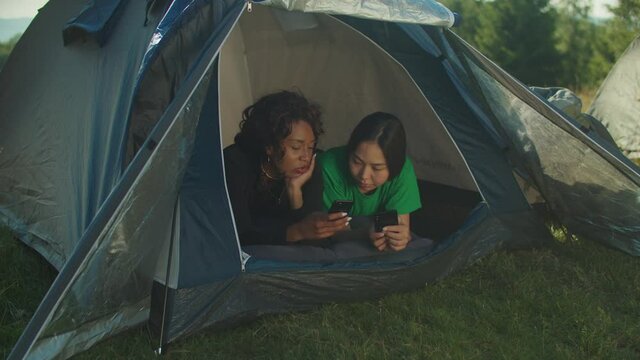 Portrait of charming relaxed multiracial women backpackers lying in camping tent, sharing and discussing social media content on cellphone while enjoying summer vacations on mountain hiking.