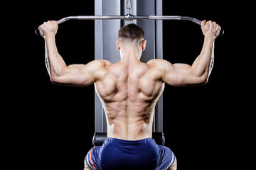 Back view of young handsome topless bodybuilder works out pulling down on cable machine in gym