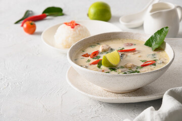 Spicy creamy coconut Tom Kha Gai soup with chicken, cherry tomato, coconut milk, kaffir, galangal, lemongrass,mushrooms on light background. Asian Thai food. Close up. Copy space.