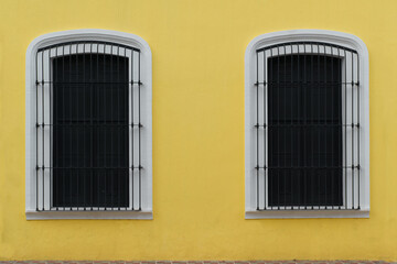 Two windows of a yellow facade and black doors with a metal grille