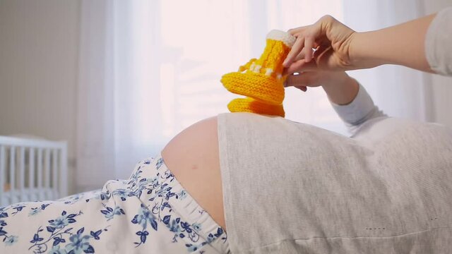 A young pregnant woman is lying on the bed, and wearing children's knitted booties on her fingers, she steps them on her tummy. Belly and hands close-up. Concept of pregnancy and motherhood .