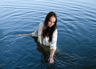 Fototapeta premium Young thoughtful brunette woman in white shirt is standing in the lake and moving hands through water.
