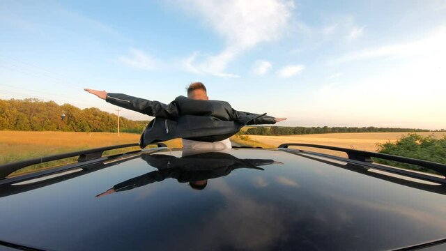 Young guy standing out of car sunroof with hands to sides and flying like plane while riding through country road. Man enjoying road trip and wind during journey. Travel or freedom concept. Back view