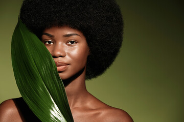 Natural skin care concept. Beauty portrait of young beautiful african american woman with posing with leaf curly hair against green exotixc plants background.