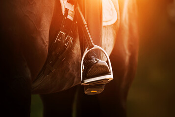 The rider's foot, sitting in the saddle on a horse, is dressed in a black boot and rests on a metal stirrup illuminated by the sun. Equestrian sports. Horse ammunition. - obrazy, fototapety, plakaty