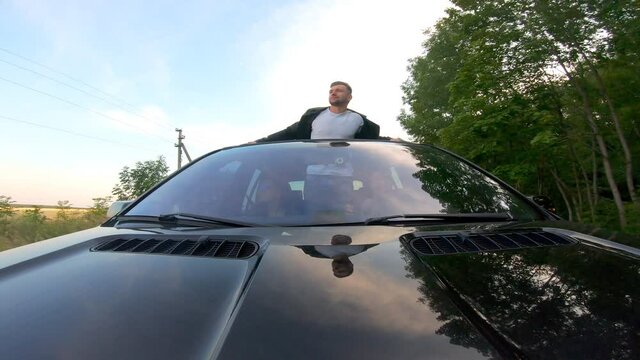 Happy funny man standing out of car sunroof and showing joyful or crazy emotions. Young guy enjoying road trip together with friends. Youth having journey by auto at sunset. Travel concept. Front view