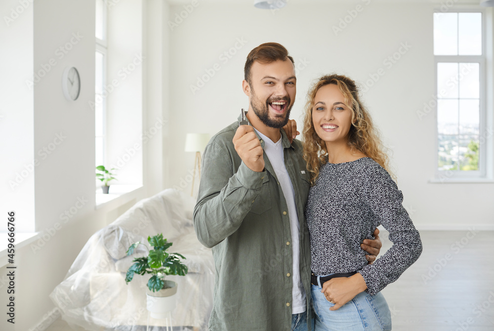 Poster Happy married couple standing against background of empty white living room in new home. Young man hugging beautiful wife, looking at camera, smiling and showing key. Real estate, buying house concept