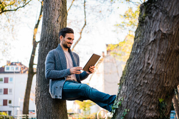 Smiling young man watching video on digital tablet while leaning on tree trunk at park