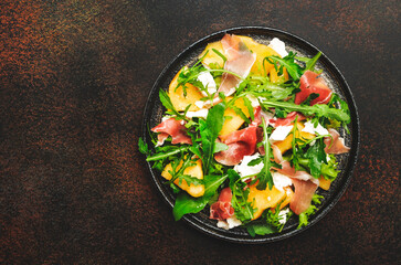 Salad with melon, jamon, goat cheese and arugula on brown background, top view, copy space