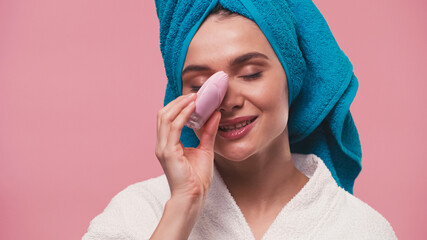 smiling woman with closed eyes cleaning face with silicone facial brush isolated on pink