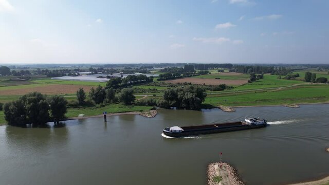Aerial pan movement rotating around and following an empty cargo vessel passing by on river IJssel near Zutphen with floodplains and wind turbines in the back. Dutch waterway inland infrastructure.