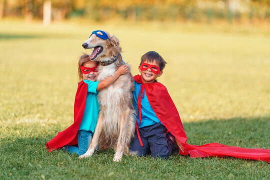little Latin American boy and a girl in a superhero costume are sitting with a large greyhound dog on a lawn in the fresh air. A child and a dog fantasize and pretend to be a superhero.