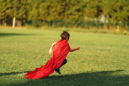 little boy in a superhero costume with a red cape runs across the lawn in the fresh air. The child fantasizes and pretends to be a superhero. A place for text or advertising. Copy space