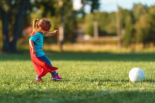 little girl child in a superhero costume is playing football in the fresh air. happy girl in red raincoat and a mask runs with a ball on the football field. Outdoor games, dynamic image