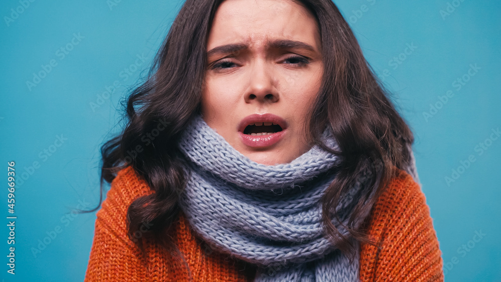 Wall mural ill woman in knitted scarf sneezing while looking at camera isolated on blue