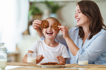 happy loving family, mom and daughter, playing sitting at table and having breakfast at home in morning. woman and girl eat oatmeal cookies and drink cow's milk, and have nice time together in kitchen