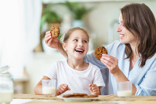 happy loving family, mom and daughter, playing sitting at table and having breakfast at home in morning. woman and girl eat oatmeal cookies and drink cow's milk, and have nice time together in kitchen