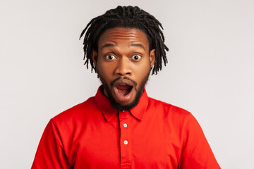 Astonished man with dreadlocks wearing red casual style T-shirt, standing with mouth open in...