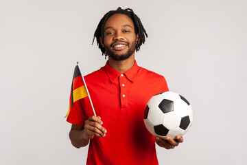 Man with toothy smile wearing red casual style T-shirt, holding flag of germany and soccer black and white classic ball and watching match. Indoor studio shot isolated on gray background. - obrazy, fototapety, plakaty