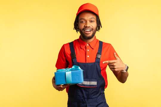 This Present For You! Happy Courier In Overalls Pointing At Blue Wrapped Box, Carrying Birthday Surprise, Delivery Service Of Purchases And Gifts. Indoor Studio Shot Isolated On Yellow Background.