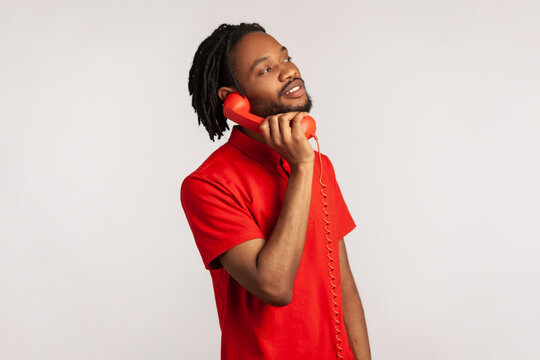 Side View Of Male With Dreadlocks And Beard Wearing Red Casual Style T-shirt, Making Calls On Landline Telephone, Looking At Camera With Toothy Smile. Indoor Studio Shot Isolated On Gray Background.