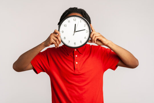 Unknown man with dreadlocks wearing red casual style T-shirt, hiding face behind big wall clock, time management, reminding of deadline. Indoor studio shot isolated on gray background.