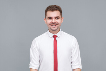Portrait of young handsome happy successful man in white shirt and tie standing and looking at camera with toothy smile. indoor isolated on gray background.