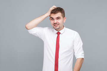 Portrait of young handsome thoughtful confused man in white shirt and tie standing scratching his head and thinking. indoor isolated on gray background.