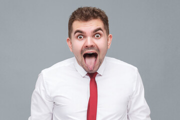 Portrait of young funny crazy man in white shirt and tie standing with big eyes and tounge out and looking at camera with crazy funny face. indoor isolated on gray background.