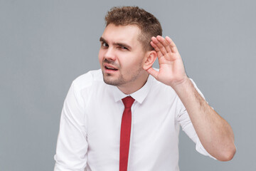 Portrait of young handsome attentive man in white shirt and tie standing with hand near ears and trying to listening. indoor isolated on gray background.