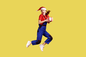 Full length portrait of delivery woman jumping high with wrapped present box, celebrating Christmas, wearing blue overalls and santa claus hat. Indoor studio shot isolated on yellow background.