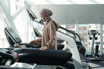 Side view shot of beautiful Muslim woman wearing hijab spending time in gym standing on treadmill...