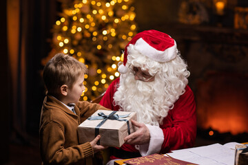 Santa Claus and little boy. Cheerful Santa is working while sitting at the table. Fireplace and Christmas Tree in the background. Christmas concept.