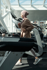 Vertical side view full shot of modern sporty Muslim woman wearing hijab running in gym using treadmill
