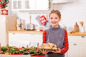 cheerful teenage girl stands near festive Christmas table and holds a dish with sweet buns in her hands.