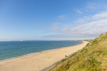 A scenic majestic view of  Bournemouth bay with sandy beach from a grassy cliff under a beautiful blue sky and some white clouds