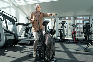 Fototapeta na wymiar Horizontal full body shot of active young adult Muslim woman wearing hijab exercising in gym using stationary bike machine