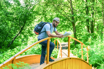 A man with a backpack in a hat stands on a wooden bridge leaning on the railing and holding a map. The traveler stopped at the bridge to read the route map