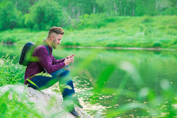 A young man, a traveler with a backpack, sits on a large stone near the river bank and looks at the phone. The tourist stopped. Copy space