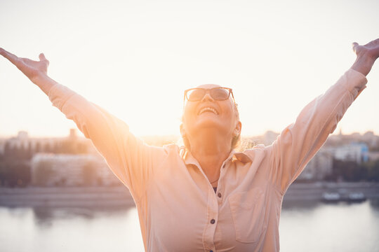 Portrait of happy attractive expressive exited mature senior woman 60 years old with hands up at sunset