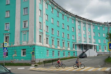  Russia, Murmansk region. Kirovsk. On a cloudy summer day, two teenage boys ride bicycles through a pedestrian crossing near the building of the hotel "Severnaya" in turquoise color