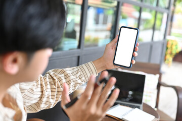 Photo of a young creative man making a video call via a white screen smartphone over an outdoor restaurant as a background.