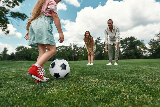Closeup of little girl kicking the ball while parents watching. Young family playing football on the grass field in the park on a summer day