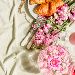 Aesthetic breakfast with croissant and pink flowers bunch on light green blanket at sunshine. Top view