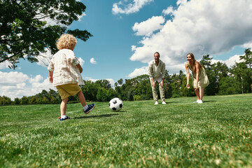 Young parents teaching their little son playing football on grass field in the park on a summer day