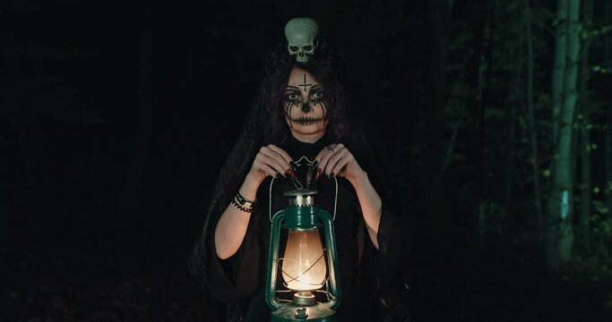 The image of a witch in a dress and a veil of make-up stands in the woods with a lantern in his hands looking terribly into the camera
