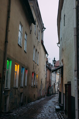 an atmospheric narrow street in the old city of Europe. Old houses, small windows with colored light. A narrow strip of cloudy sky is visible. The houses are three-storied