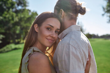 Portrait of pretty young woman smiling, embracing her husband, posing together outdoors while standing in the park