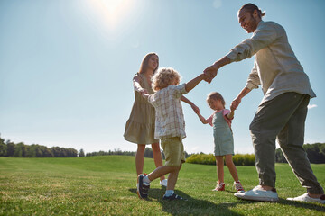 Full length shot of young parents holding hands in circle with their two little kids, boy and girl in green park on a summer day